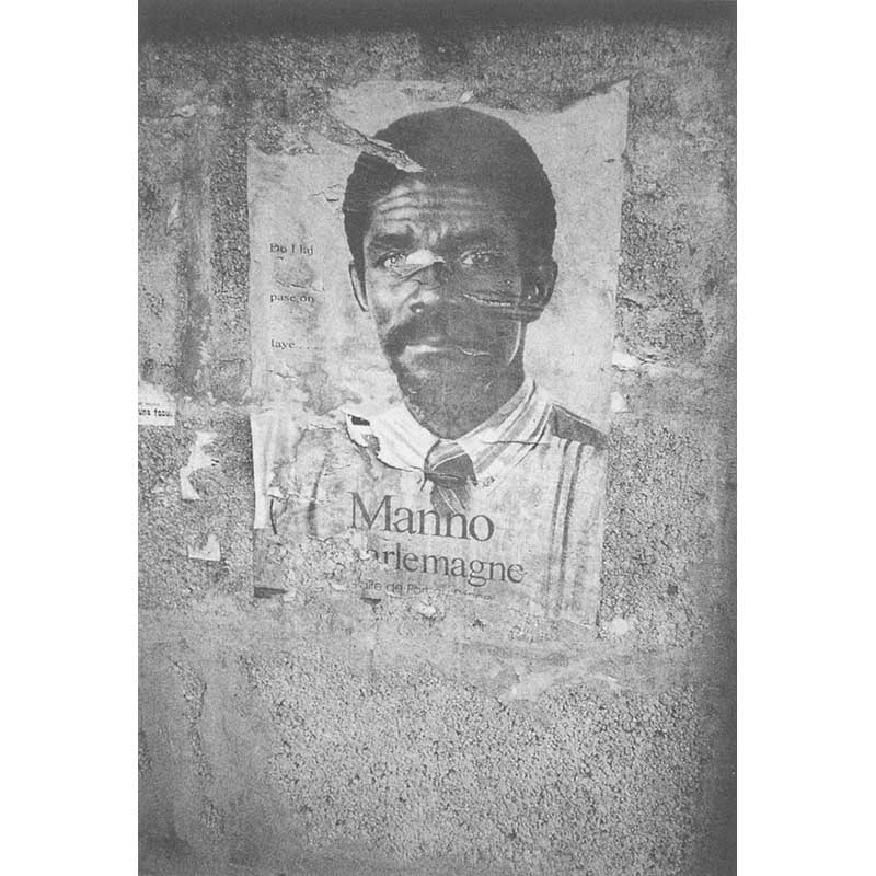 Manno Charlemagne's face appears on a torn poster on the wall.